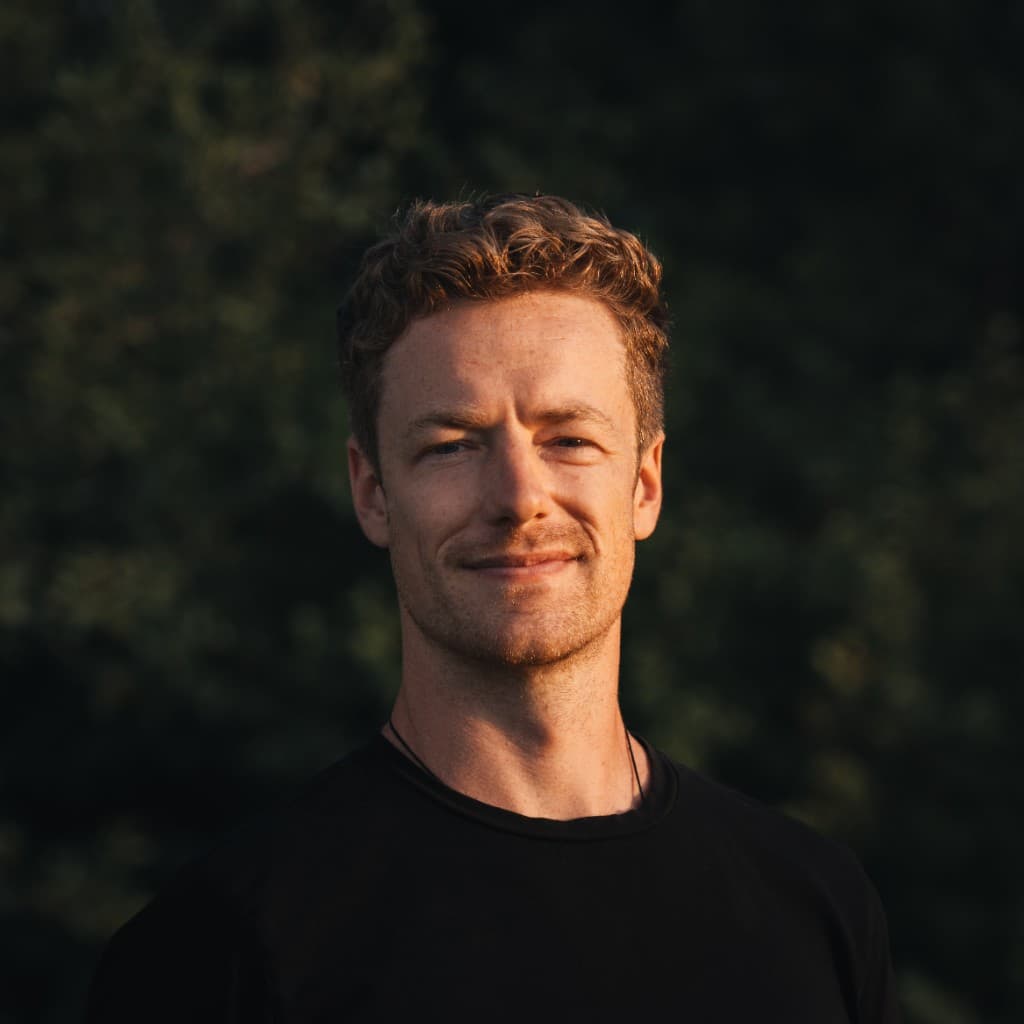 Professional headshot of Matt Hennessy, smiling, lit by warm sunlight against a blurred dark green background.
