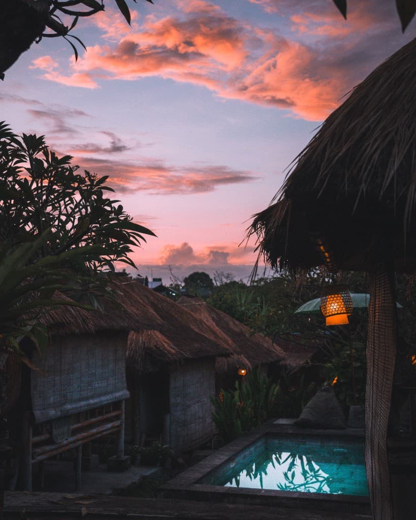 Thatched-roof huts and a glowing pool under a pink and purple tropical sunset.