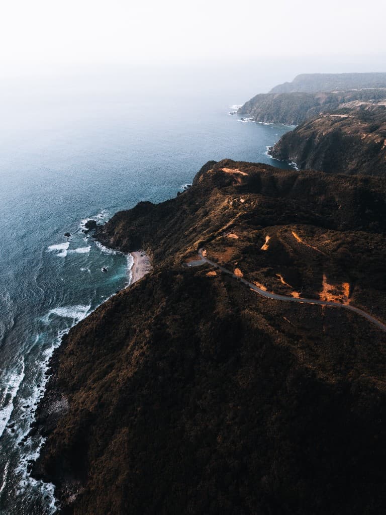 Aerial view of dramatic coastal cliffs and a winding road above crashing waves.