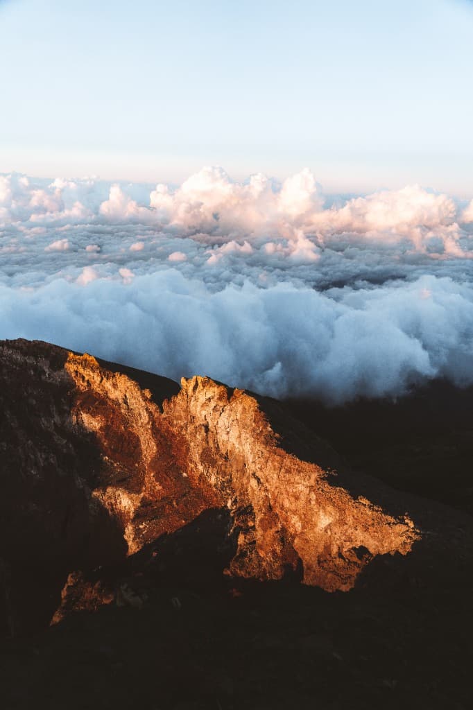 Sunlit volcanic rock on a crater rim with a sea of clouds below.