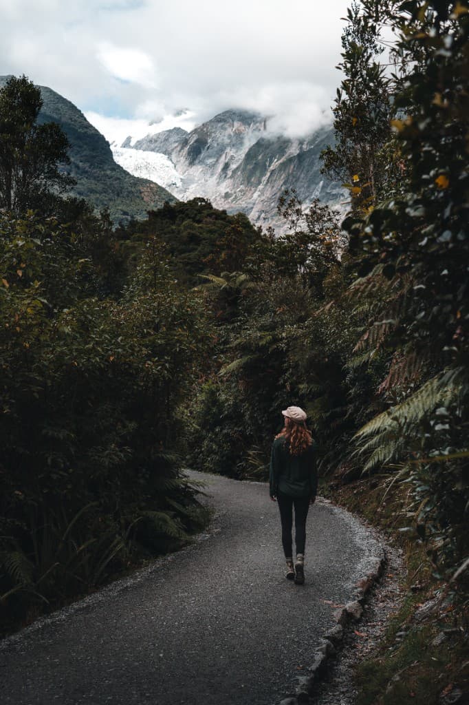 A person walking a gravel path through lush rainforest toward a distant glacier.