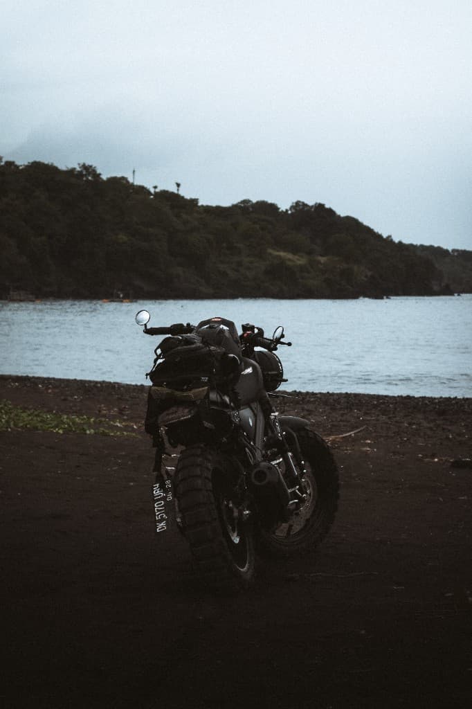 A motorcycle parked on a dark volcanic beach with hills across the water.
