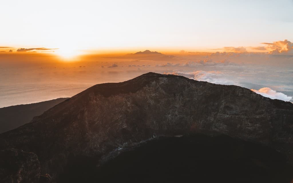 Sunrise above the clouds from a volcanic summit with a distant peak on the horizon.