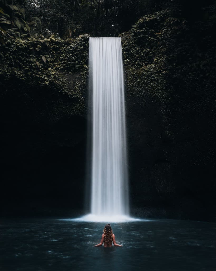 A person waist-deep in water facing a tall, smooth waterfall surrounded by moss-covered rock.