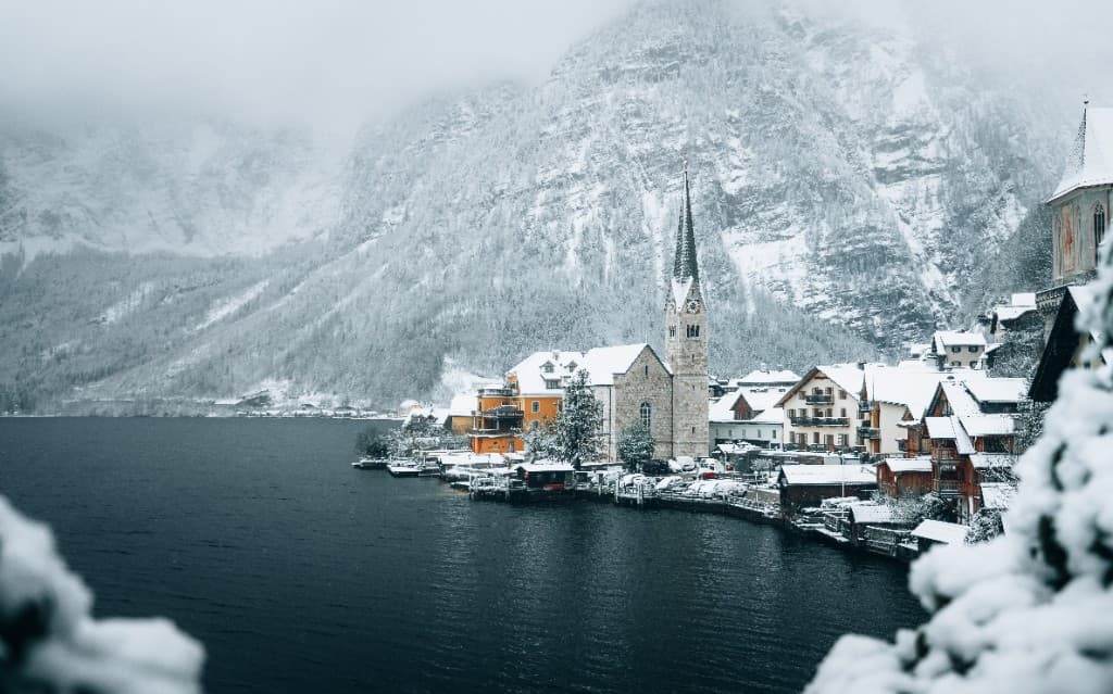 A snowy lakeside village with a church spire beneath fog-covered alpine mountains.